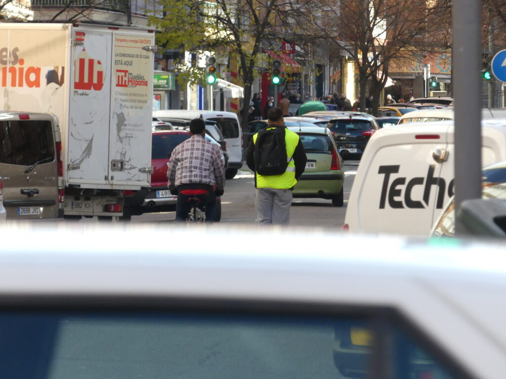 Jóvenes en bicicleta y patinete por una carretera llena de coches.